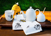 glossy white bone china tea pot in shape of beehive with long curled spout beside glossy white honey pot with dipper, white mug with yellow honey bee, felt beehive and bee tea towel on dark wood table with green grass in background