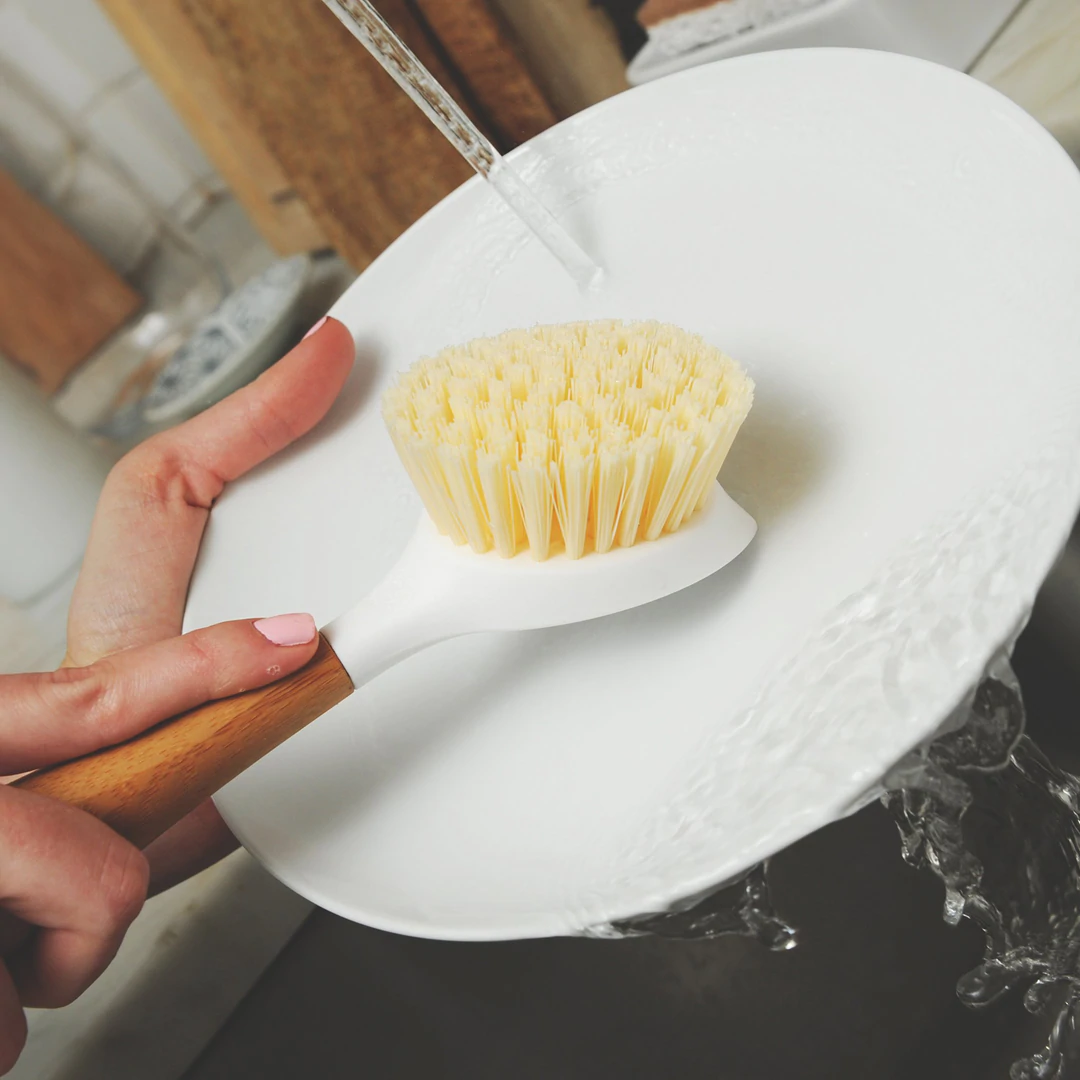 Caucasian person scraping dish with back end of dish brush under running water by full circle