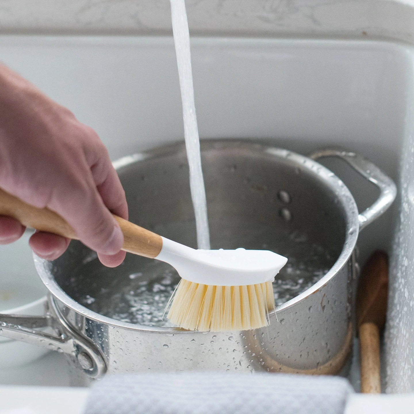 Caucasian person washing silver pot under running water with bamboo dish brush by full circle