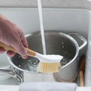 Caucasian person washing silver pot under running water with bamboo dish brush by full circle