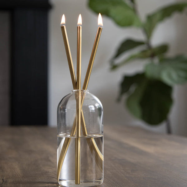 Clear glass diffuser with gold CANDLE sticks on a wooden surface, blurred plant in the background