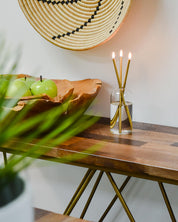 Wooden table with a bowl of green apples, a glass vase with candles, and a woven basket on a white wall.