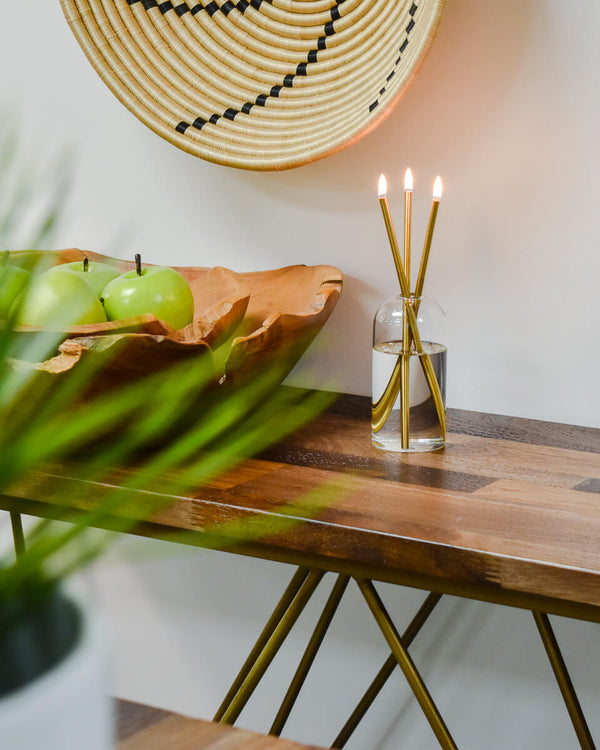 Wooden table with a bowl of green apples, a glass vase with candles, and a woven basket on a white wall.