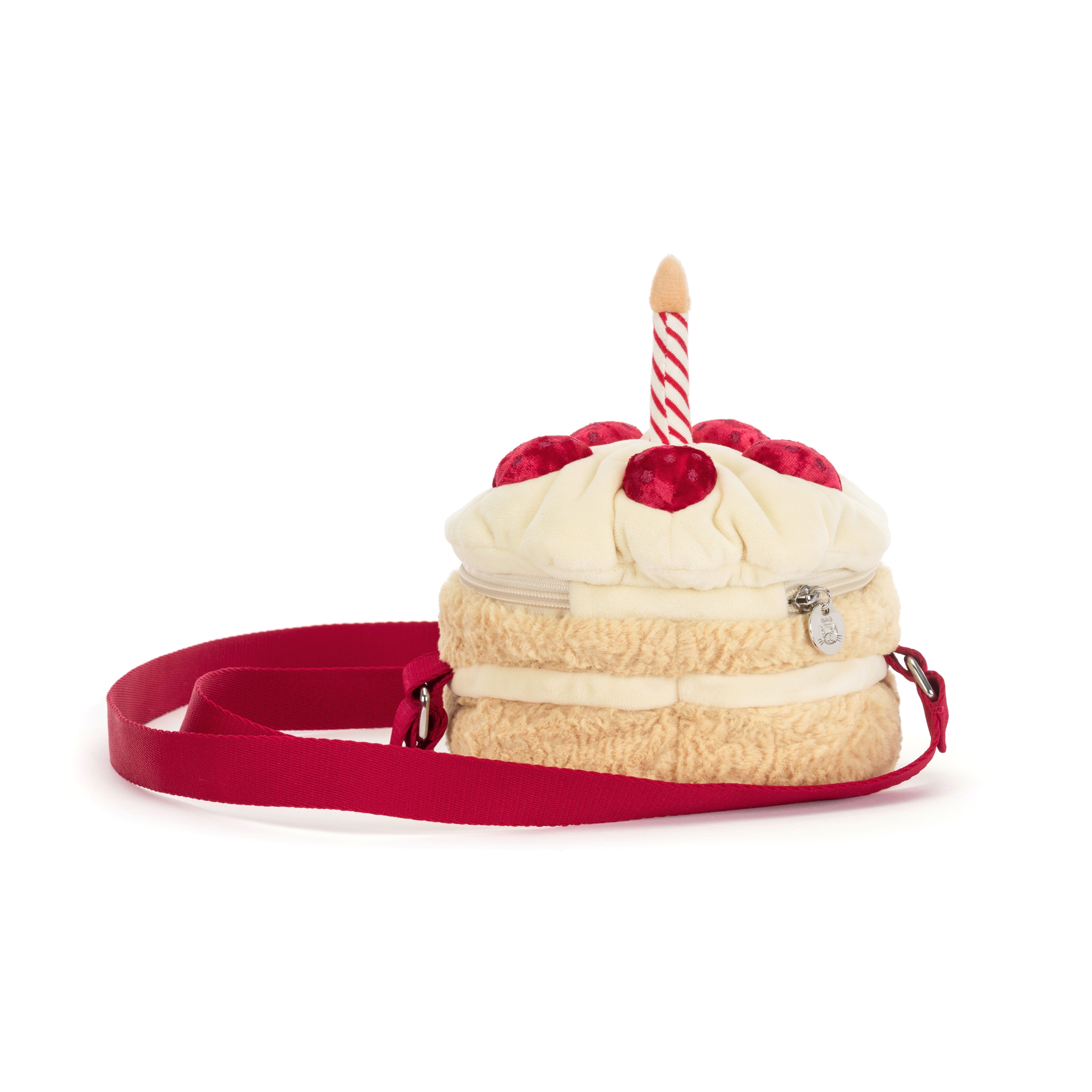 A plush cake-shaped shoulder bag with cream-colored faux frosting, red strawberries, a red strap, and a single red-and-white striped candle on top, shown on a plain white background.