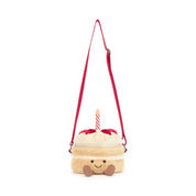 A plush crossbody bag shaped like a cute cream-colored cake with red strawberries and a red and white stripe candle, featuring a red shoulder strap and a smiling face on a white background.