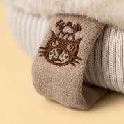 close up of the tag on A plush winter mitten jellycat toy in a warm taupe-brown fabric with cream sherpa trim, featuring a simple embroidered smiley face 