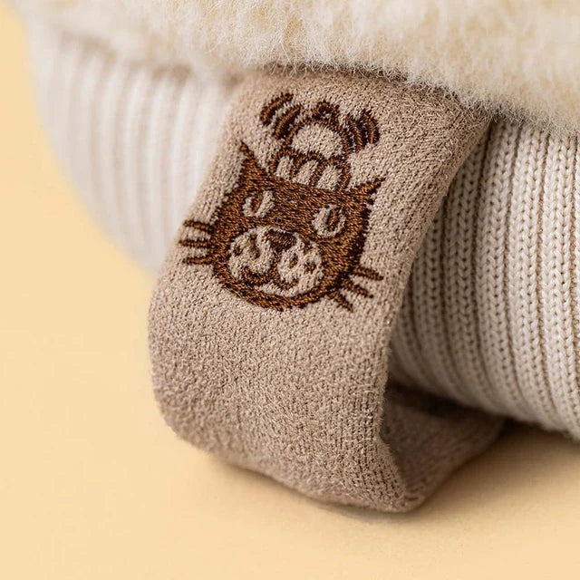 close up of the tag on A plush winter mitten jellycat toy in a warm taupe-brown fabric with cream sherpa trim, featuring a simple embroidered smiley face 