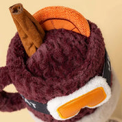 top view of A jellycat brand plush toy shaped like a  mug of mulled spiced drink with a smiling face, wearing orange ski goggles and brown boots, on a solid white background.