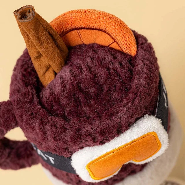 top view of A jellycat brand plush toy shaped like a  mug of mulled spiced drink with a smiling face, wearing orange ski goggles and brown boots, on a solid white background.