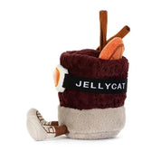 sideview of A jellycat brand plush toy shaped like a  mug of mulled spiced drink with a smiling face, wearing orange ski goggles and brown boots, on a solid white background.