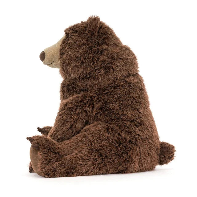 sideview of A large jellycat brown plush stuffed bear toy sitting upright on a clean white background.