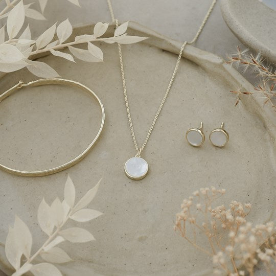 Gold jewelry set featuring a necklace with a round white stone pendant, matching stud earrings, and a gold bangle, arranged on a textured beige tray with dried botanicals.