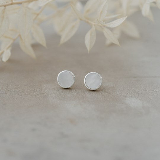 A pair of minimalist silver stud earrings with flat circular faces, resting on a neutral textured surface with soft, out-of-focus dried leaves in the background.