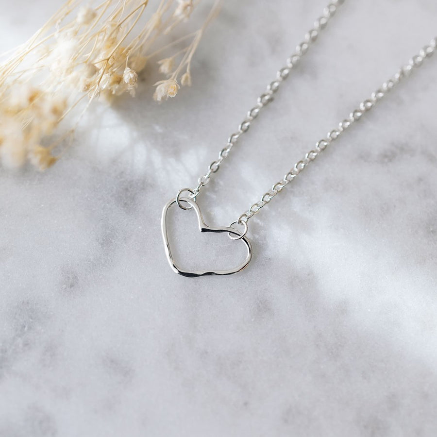 A delicate silver chain necklace with an open heart-shaped pendant, displayed on a light gray marble surface with dried flowers in the corner.