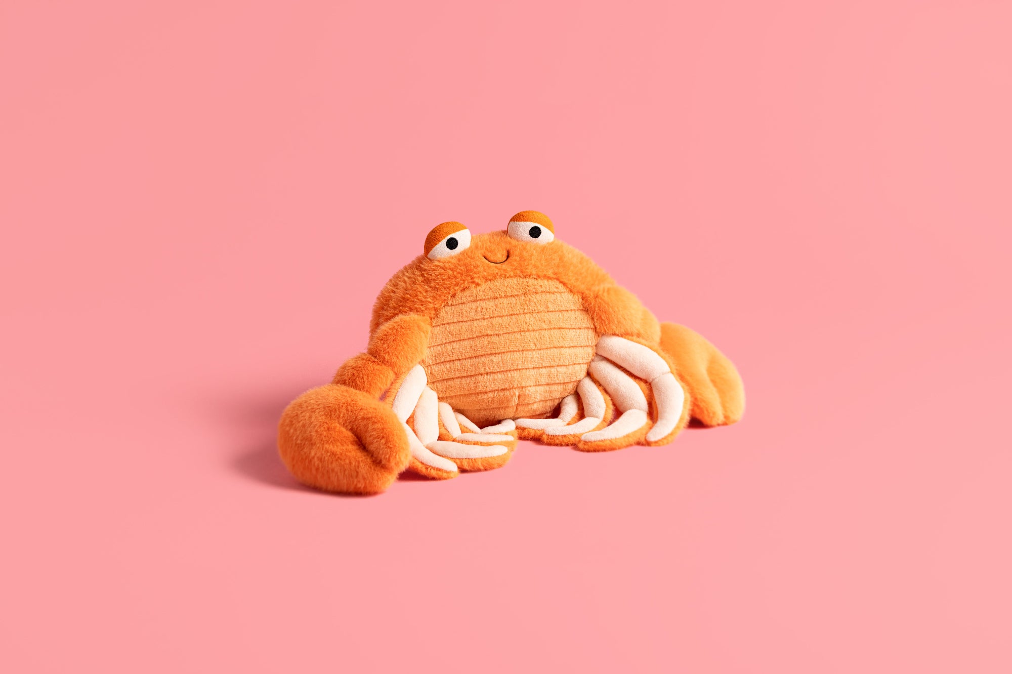 Plush crab jellycat with orange and beige colours on a pink background