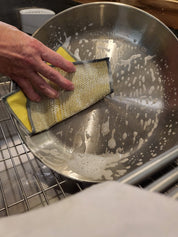 Person cleaning a metal pot with a jude's cleaning scrubbie in a kitchen sink.