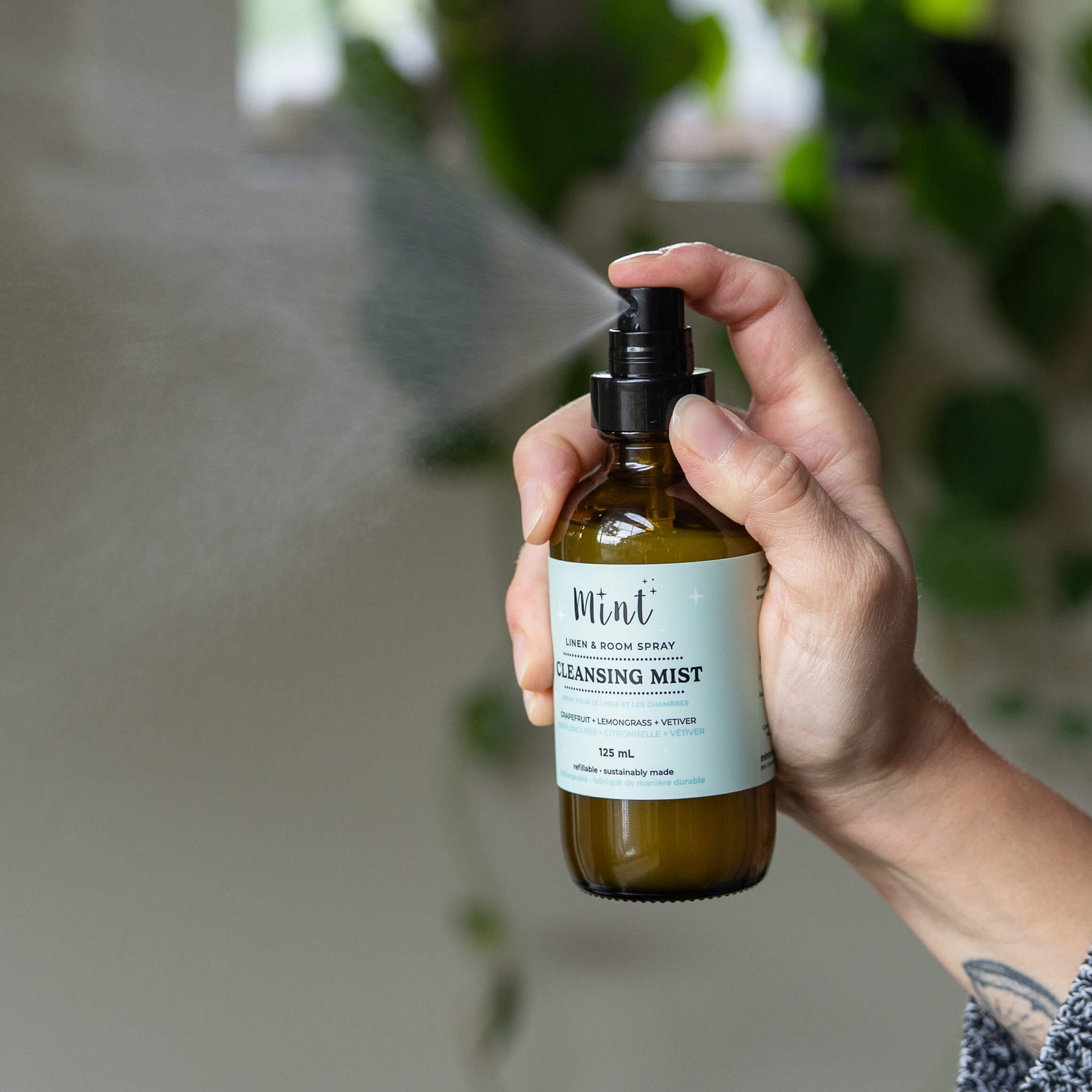 Hand holding a bottle of cleansing mist with a blurred green plant background