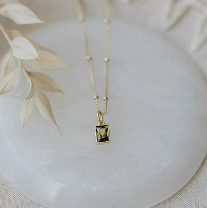 A gold-colored necklace with a moss green crystal pendant on a white marble surface, surrounded by decorative dry leaves.