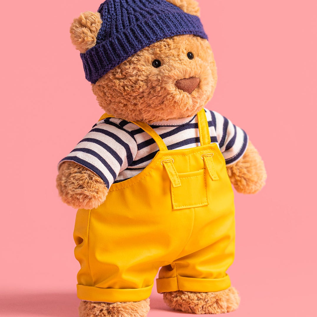 jellycat bartholomew Teddy bear dressed as a sailor wearing a navy hat, striped shirt, and yellow overalls on a pink background