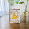 white greeting card with smiling hat wearing cheese illustration with gold lettering that reads "you get better with age! happy birthday" standing on wood table