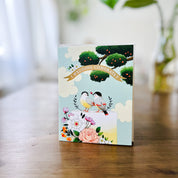 sky blue greeting card with illustration of apple tree and wedding cake with flowers and birds sitting on top with gold banner that reads "wedding congrats" standing on wood table