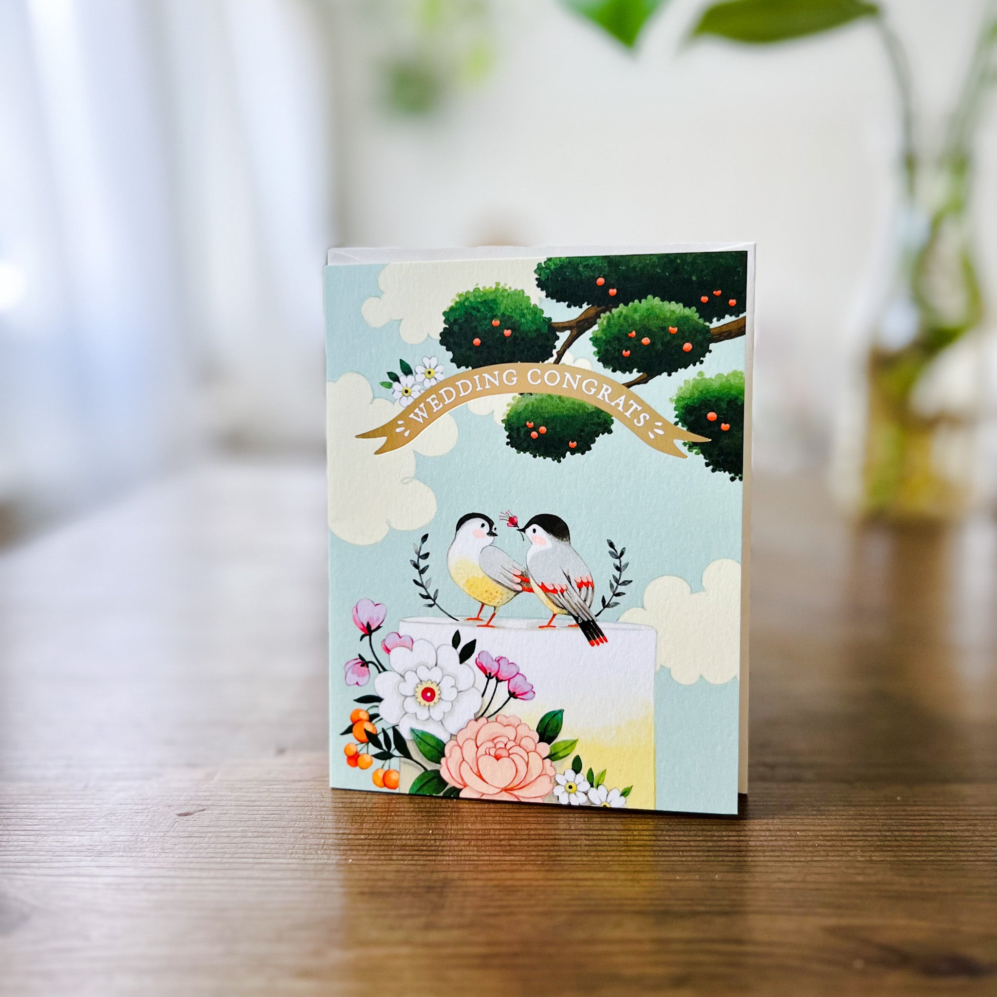 sky blue greeting card with illustration of apple tree and wedding cake with flowers and birds sitting on top with gold banner that reads "wedding congrats" standing on wood table
