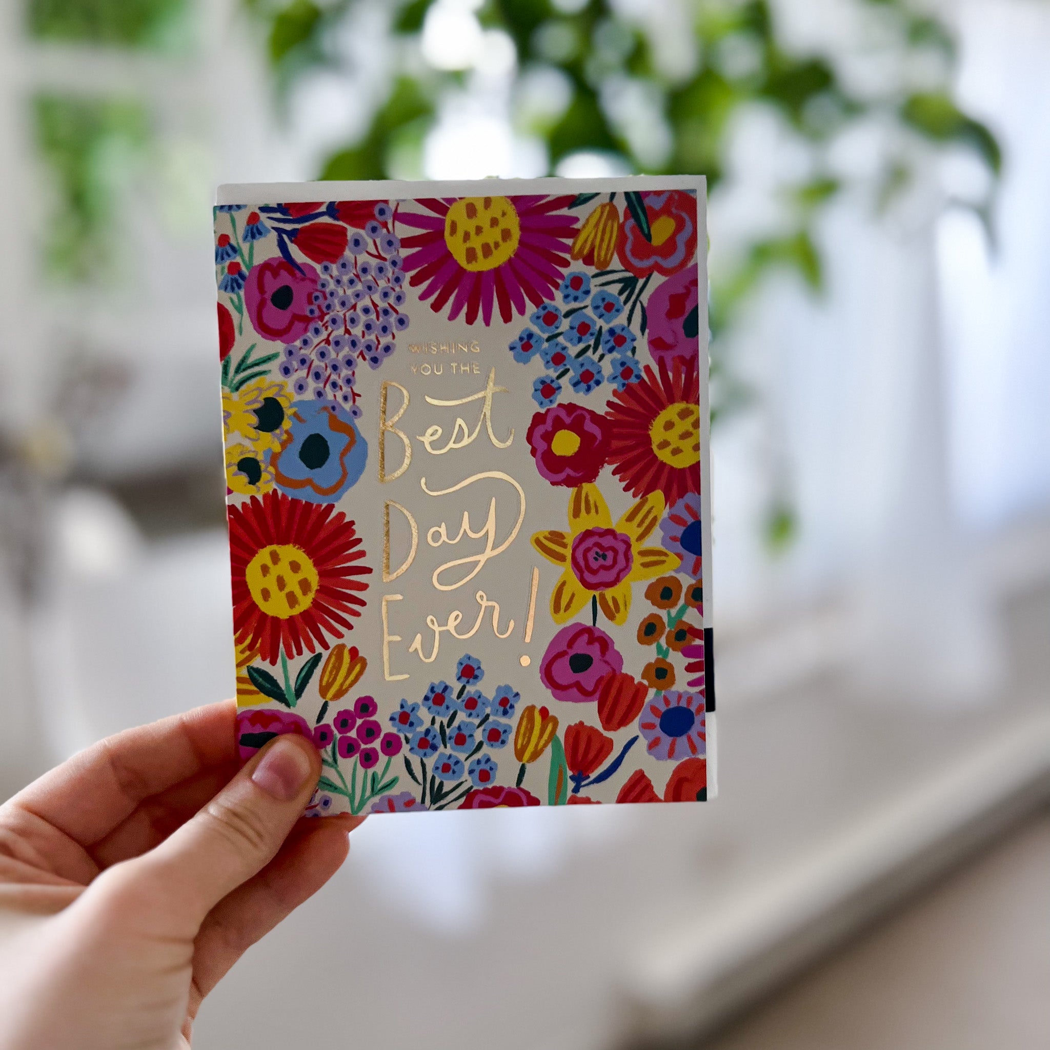 Caucasian hand holding brightly coloured floral greeting card with words that read "wishing you the best day ever!" in shiny gold lettering