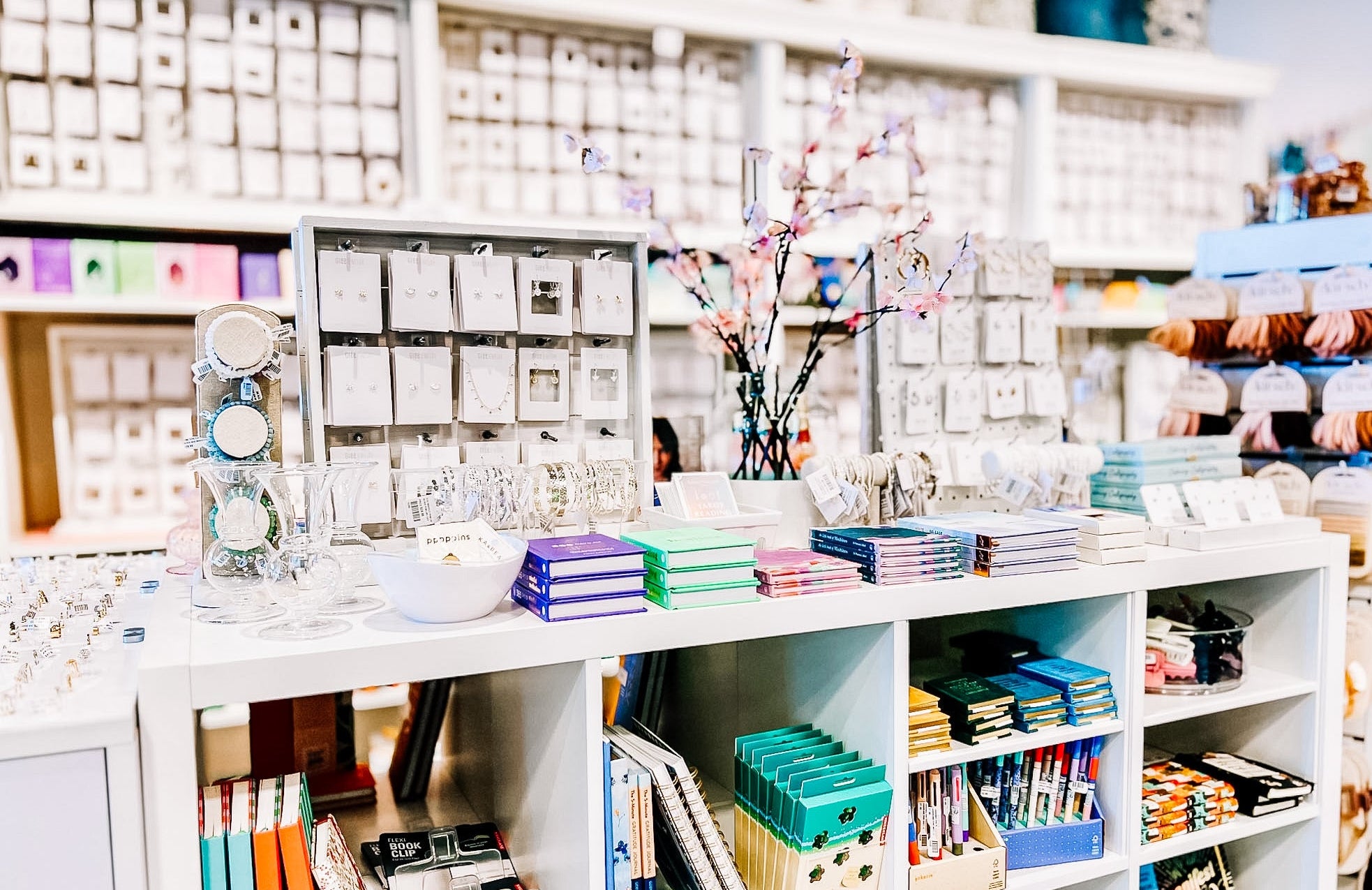 bright coloured stationary on white table and shelving with cherry blossom branches and jewelry on table and more shelving of jewelry behind 