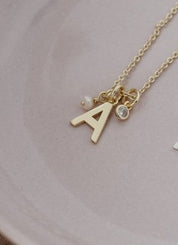 A delicate gold initial necklace with the letter 'A' and small charms, resting on a light pink ceramic dish.