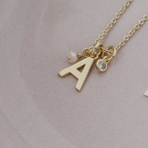 A delicate gold initial necklace with the letter 'A' and small charms, resting on a light pink ceramic dish.