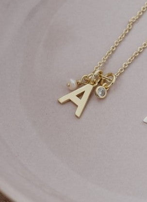 A delicate gold initial necklace with the letter 'A' and small charms, resting on a light pink ceramic dish.