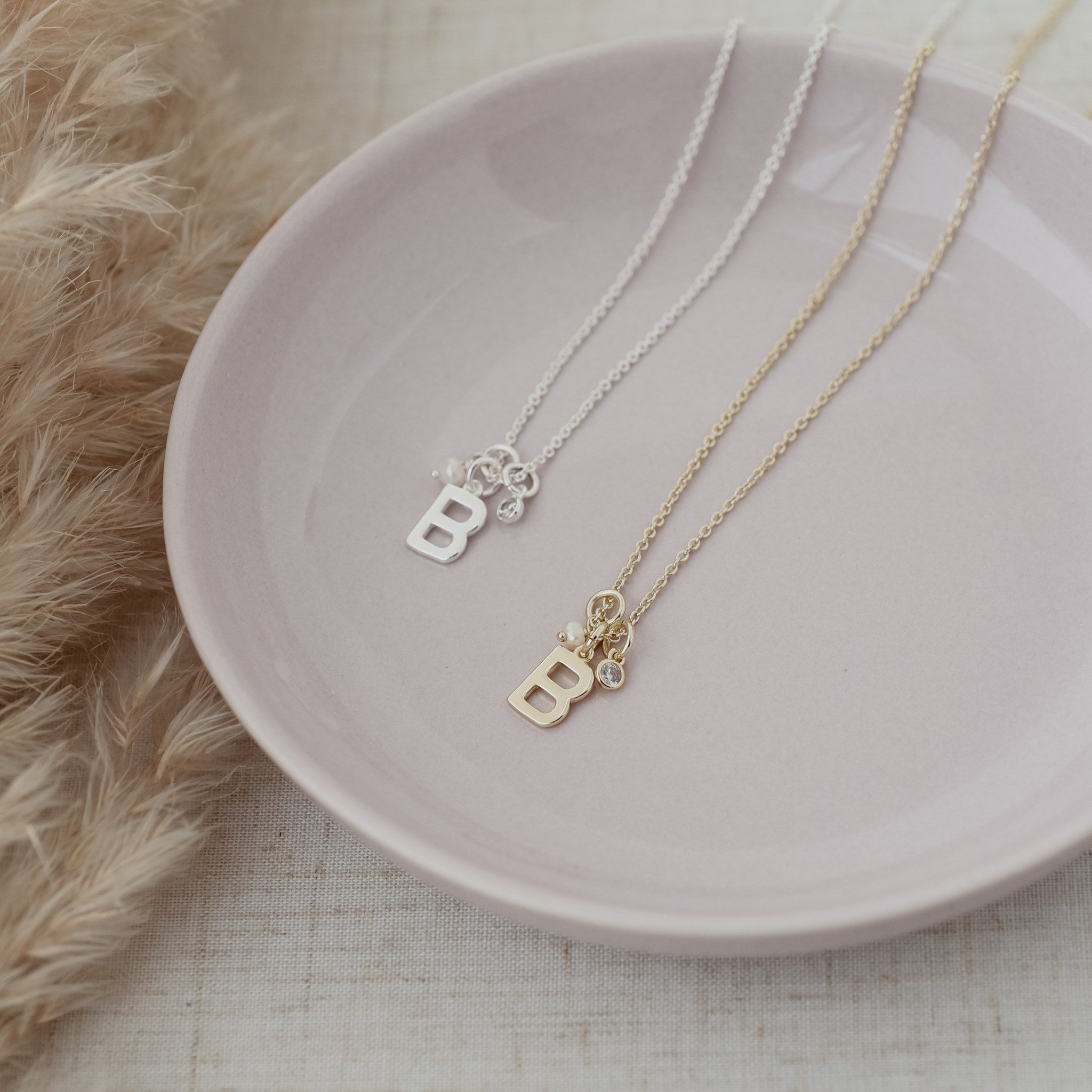 Two delicate initial necklaces with letter B pendants, one silver and one gold, displayed on a light pink ceramic plate with dried pampas grass on a neutral textured surface.