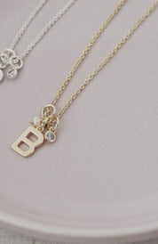 Two delicate necklaces, one gold and one silver, displayed on a pale pink ceramic plate. The gold necklace features a small letter 'B' pendant with a tiny clear crystal accent, while the silver necklace has a small floral pendant.