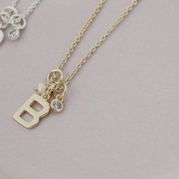 Two delicate necklaces, one gold and one silver, displayed on a pale pink ceramic plate. The gold necklace features a small letter 'B' pendant with a tiny clear crystal accent, while the silver necklace has a small floral pendant.