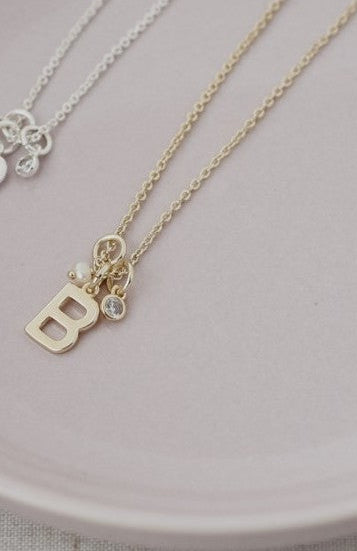 Two delicate necklaces, one gold and one silver, displayed on a pale pink ceramic plate. The gold necklace features a small letter 'B' pendant with a tiny clear crystal accent, while the silver necklace has a small floral pendant.