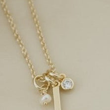 A delicate gold necklace with a letter 'L' initial pendant, a small clear crystal charm, and a tiny pearl charm, displayed on a warm cream-colored ceramic plate.