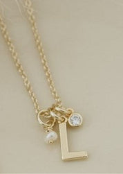 A delicate gold necklace with a letter 'L' initial pendant, a small clear crystal charm, and a tiny pearl charm, displayed on a warm cream-colored ceramic plate.