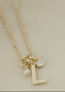 A delicate gold necklace with a letter 'L' initial pendant, a small clear crystal charm, and a tiny pearl charm, displayed on a warm cream-colored ceramic plate.