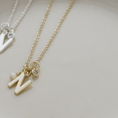 Two delicate initial necklaces with letter 'M' pendants, one in gold and one in silver, displayed on a white ceramic plate.