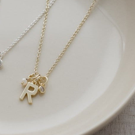 A delicate gold initial necklace with an 'R' charm and a small matching charm, displayed on a light beige ceramic plate.
