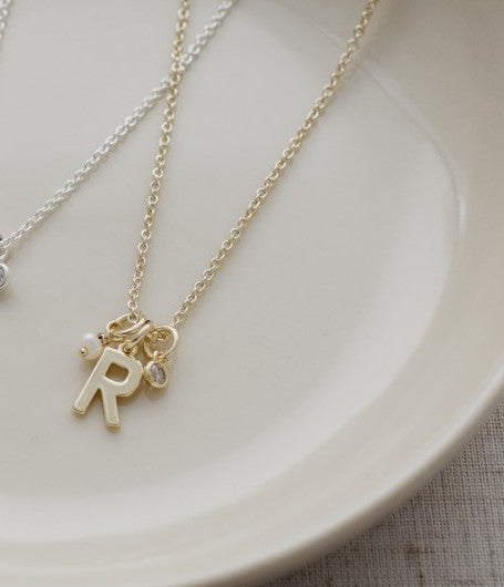 A delicate gold initial necklace with an 'R' charm and a small matching charm, displayed on a light beige ceramic plate.