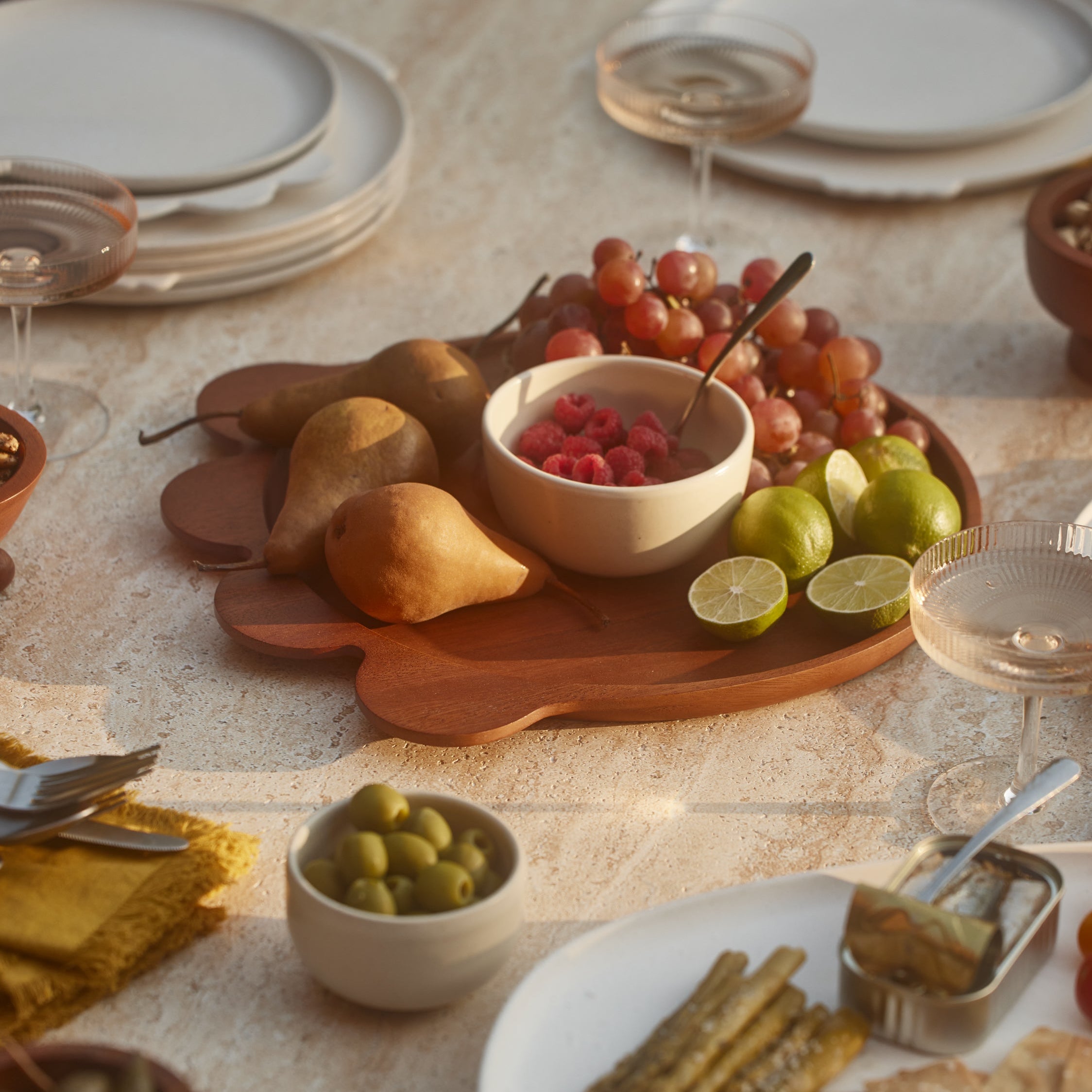 Dining table set with plates, bowls of fruit and snacks, and glasses on a neutral background.