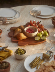 Dining table set with plates, bowls of fruit and snacks, and glasses on a neutral background.