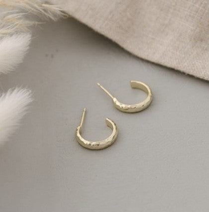 A pair of small gold huggie hoop earrings with a textured finish, displayed on a neutral gray surface with decorative white pampas grass and a beige linen cloth.