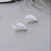 Pair of gold and silver MUSSEL SHELL-shaped earrings on a light gray surface with seashells in the background.