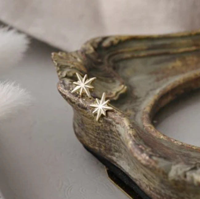 A pair of gold stud earrings with a star design, placed on a textured surface with a white furry object to the left.