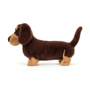 Side view of plush sausage dog toy with dark brown fur, beige feet and snout, with black nose and eyes on a white background.