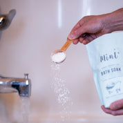 Person measuring bath salt with a wooden scoop near a container of bath soap.