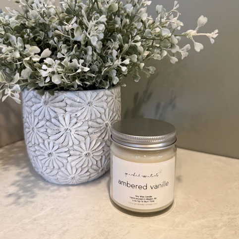 Candle labeled 'ambered vanille' next to a decorative plant on a textured surface.