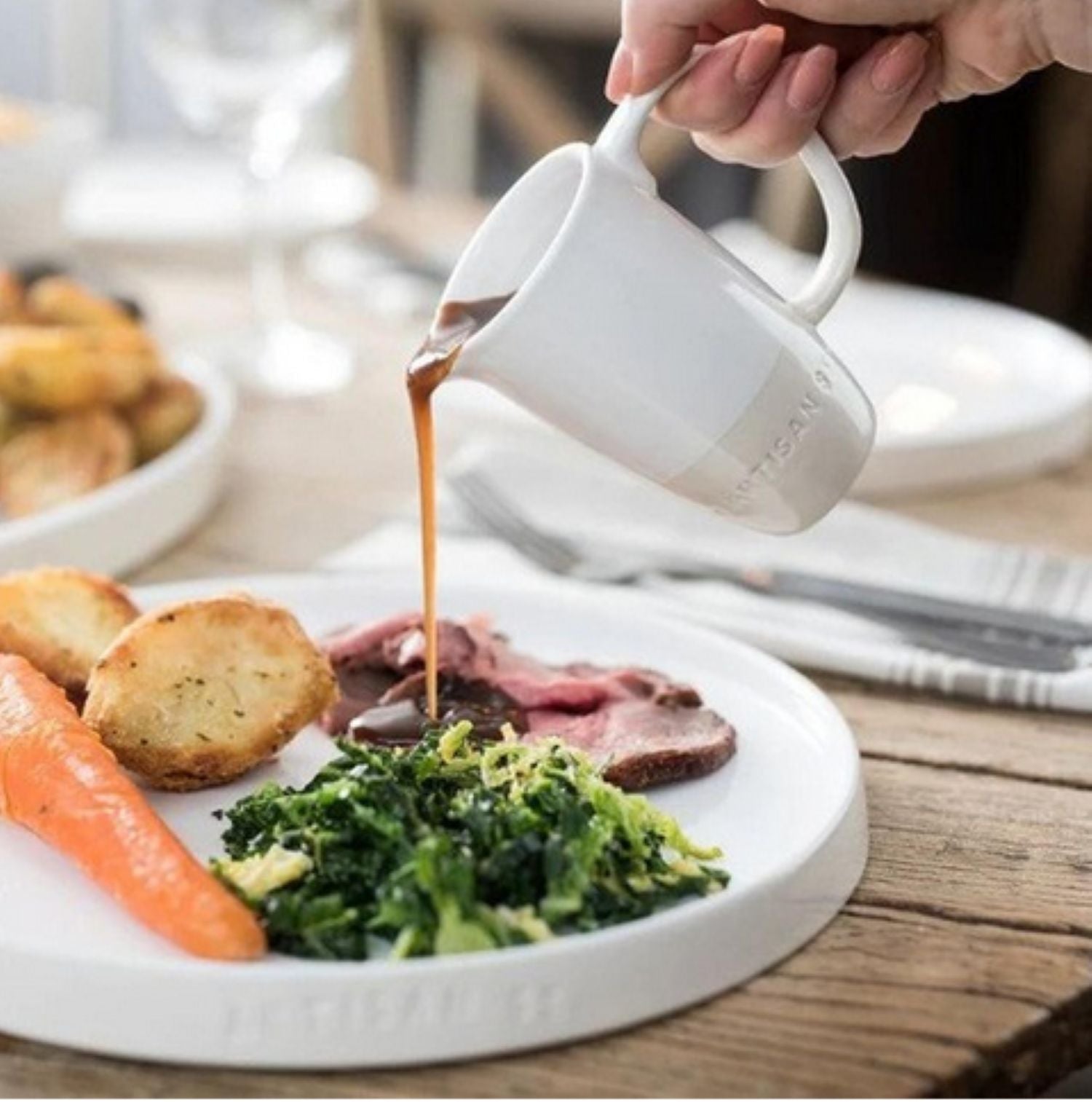 Person pouring gravy from a white pitcher onto a plate of food on a wooden table.